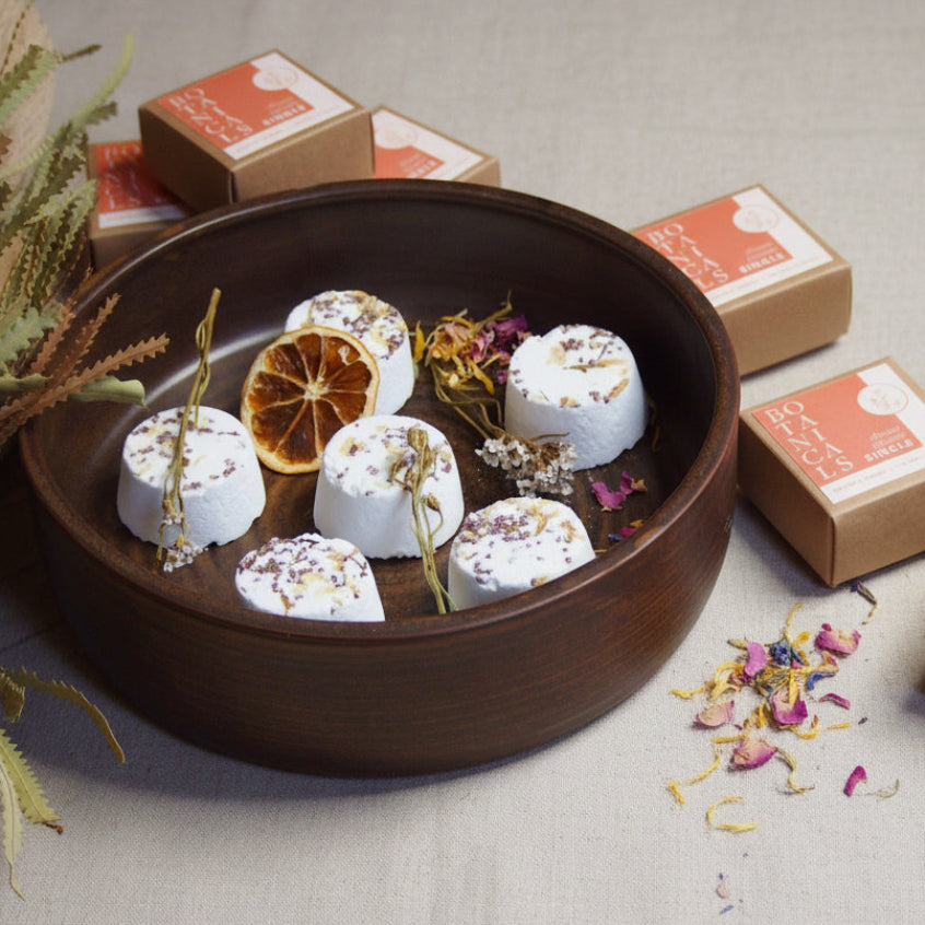 An array of single shower steamers with various botanical ingredients, placed on a wooden surface with decorative flowers and citrus fruits around them. The steamers are handmade and displayed in a natural setting.
