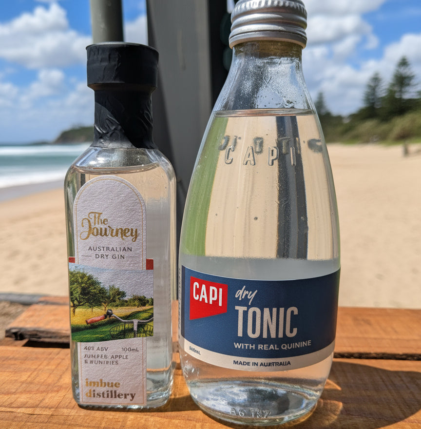 Two bottles of alcohol, one labeled 'The Journey' and the other 'Capi dry Tonic', on a wooden surface with a garden background.