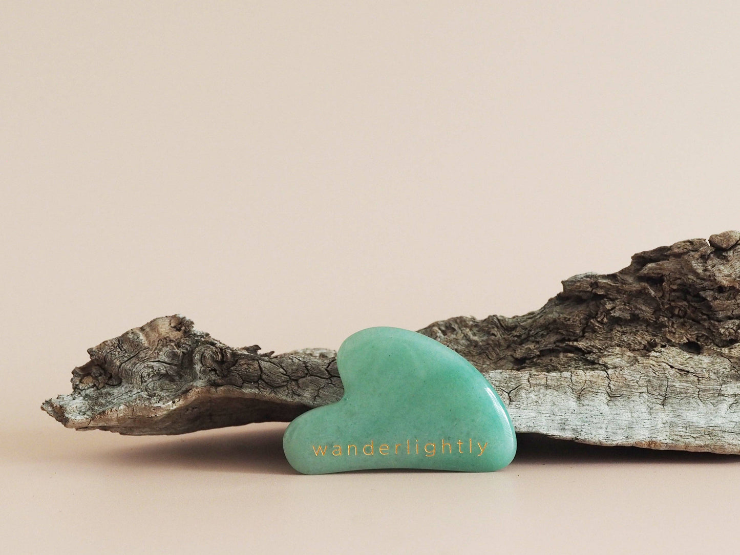 A green Jade Gua Sha tool placed on a piece of driftwood against a neutral background.