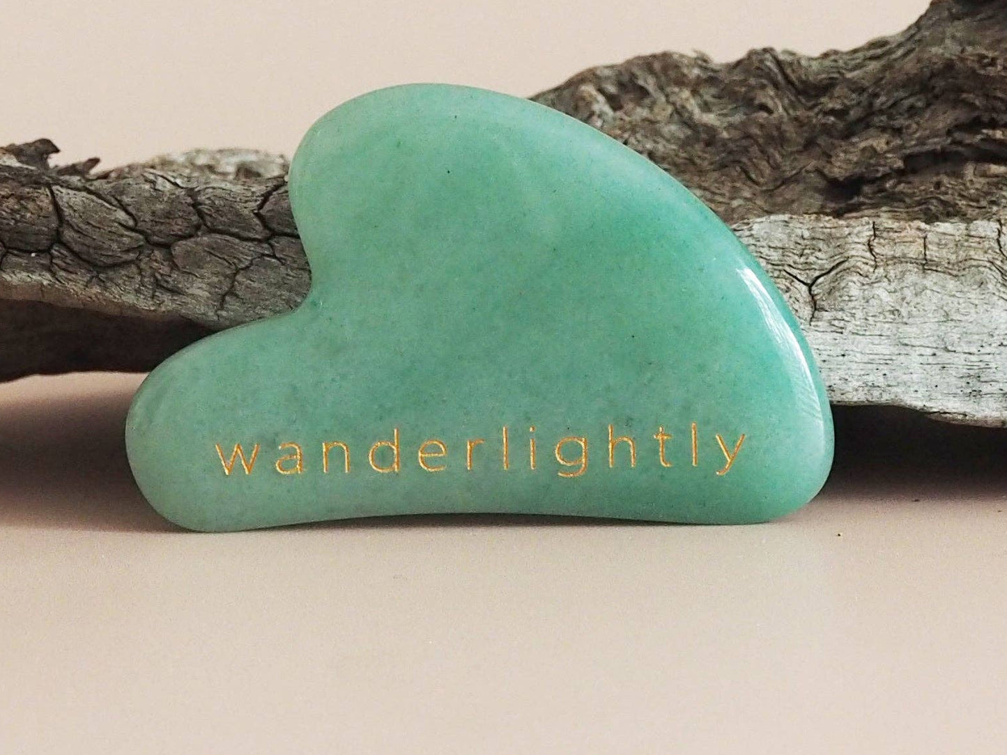 A green Jade Gua Sha tool placed on a piece of driftwood against a neutral background.