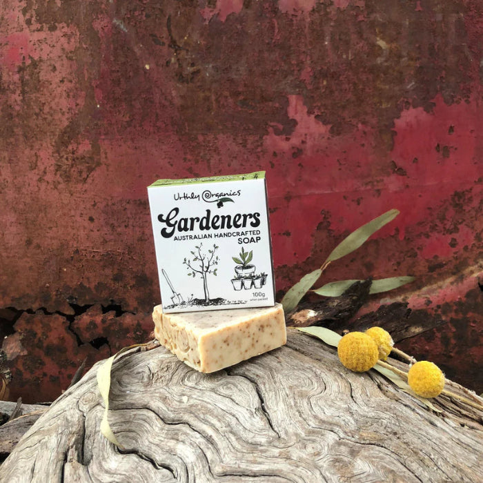 A bar of soap with a label that reads 'GARDENERS SOAP SPECIALTY SOAP' placed on a rusty metal surface with a few dried flowers and eucalyptus leaves scattered around.