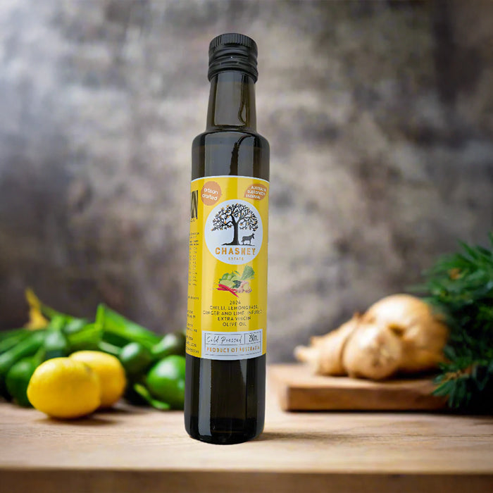 A bottle of Chilli Lemongrass Ginger and Lime Infused Extra Virgin Olive Oil placed on a wooden surface with a background of lemon slices, ginger root, and pine branches.