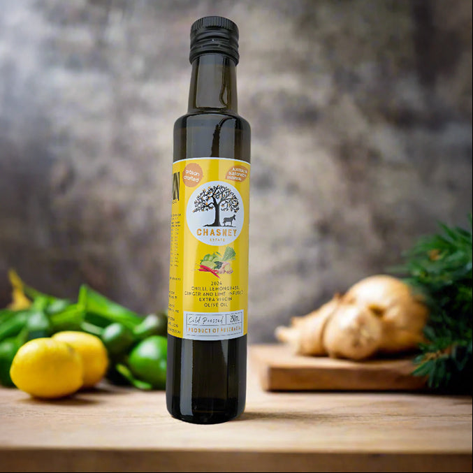 A bottle of Chilli Lemongrass Ginger and Lime Infused Extra Virgin Olive Oil placed on a wooden surface with a background of lemon slices, ginger root, and pine branches.