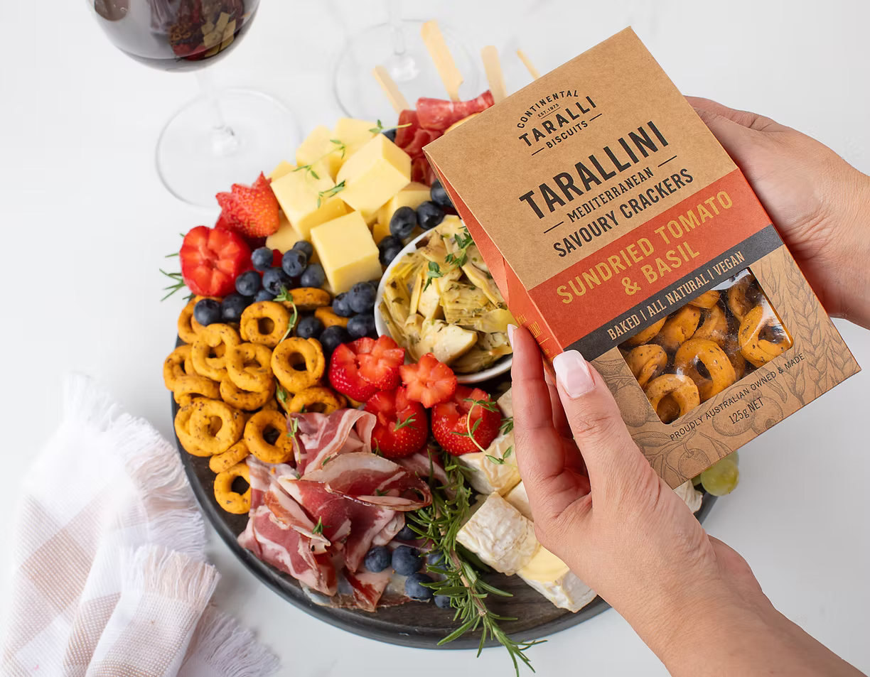 Hand holding a box of Tarallini with a charcuterie board in the background