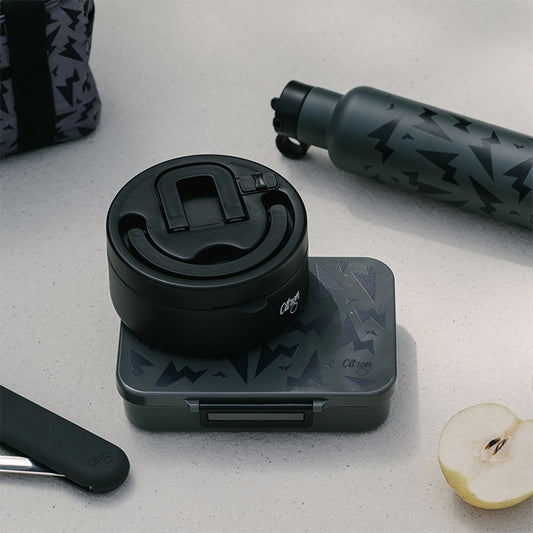 A black 400ml stainless steel thermal food jar with a geometric pattern on the lid, placed on a matching black stand, next to a gray water bottle and a apple.
