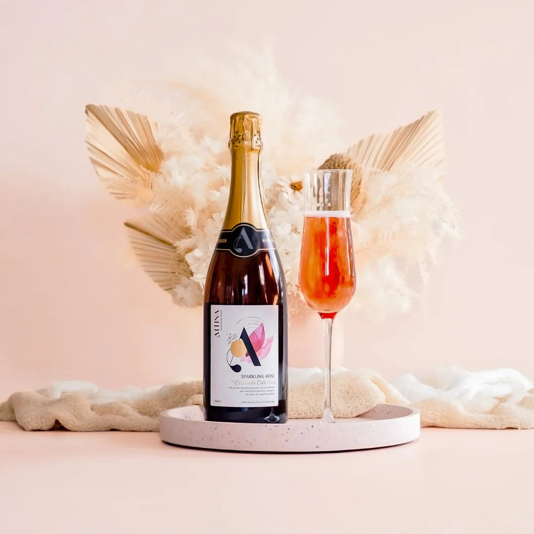 Bottle of sparkling wine with a glass of pink cocktail on a decorative stand against a soft pink background.