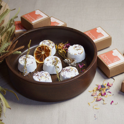 An array of single shower steamers with various botanical ingredients, placed on a wooden surface with decorative flowers and citrus fruits around them. The steamers are handmade and displayed in a natural setting.