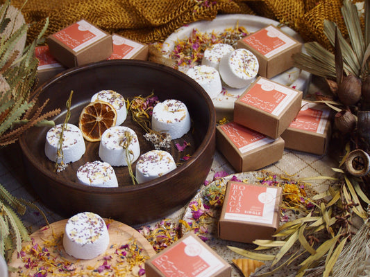 An array of single shower steamers with various botanical ingredients, placed on a wooden surface with decorative flowers and citrus fruits around them. The steamers are handmade and displayed in a natural setting.
