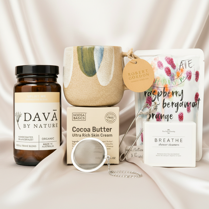 Collection of self-care products including organic tea bottle, body cream, mug, chocolate and shower steamer.