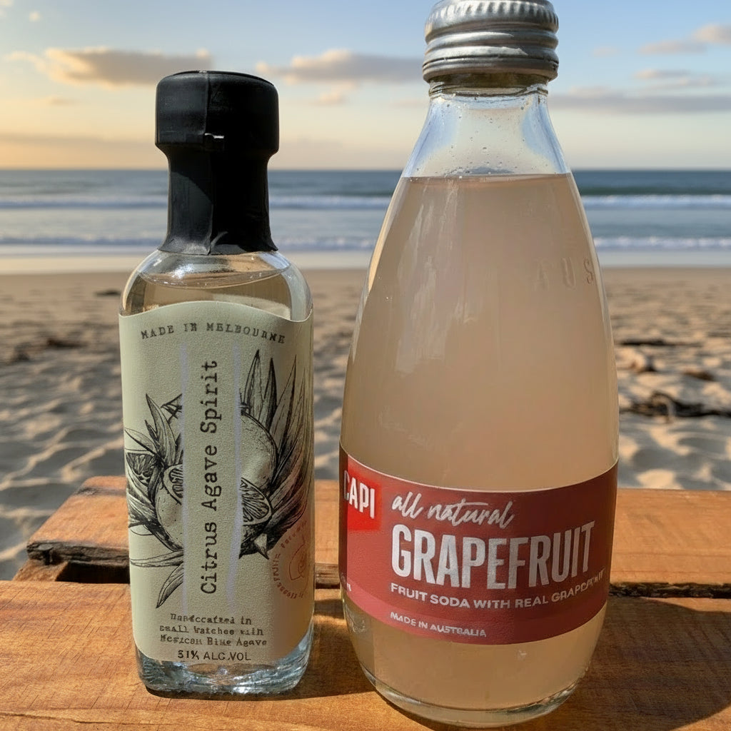 Two bottles, one labeled 'Citrus Agave Spirit' and the other 'Grapefruit Soda', on a wooden surface with a grassy area in the background.