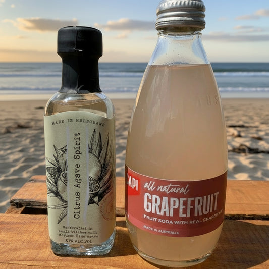 Two bottles, one labeled 'Citrus Agave Spirit' and the other 'Grapefruit Soda', on a wooden surface with a grassy area in the background.