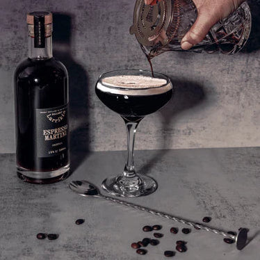Bottle of espresso martini and a glass being filled with a dark liquid on a gray surface.