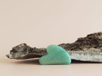A green Jade Gua Sha tool placed on a piece of driftwood against a neutral background.