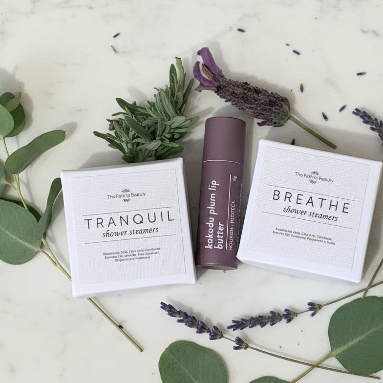Cosmetic products including 'Tranquil' and 'Breathe' shower steamers and 'Kakadu Plum Lip Butter' on a light background with lavender and eucalyptus leaves.