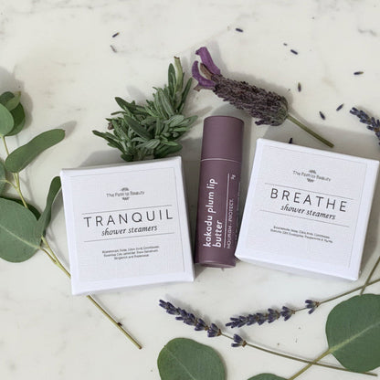 Cosmetic products including 'Tranquil' and 'Breathe' shower steamers and 'Kakadu Plum Lip Butter' on a light background with lavender and eucalyptus leaves.