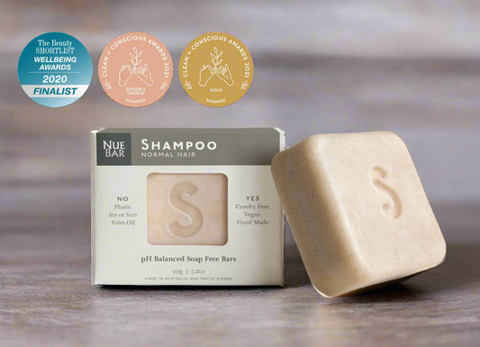 A solid shampoo bar alongside its packaging box, with badges indicating awards won. The packaging is labeled 'Shampoo for Normal Hair' and mentions organic and fair-trade ingredients.