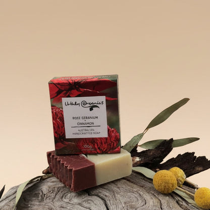 A bar of handcrafted soap next to its packaging, with a red background and a few dried flowers on a wooden surface.