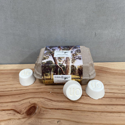 A carton with three white toilet bomb tablets placed on a wooden surface. The carton has a window displaying a natural scene with trees.