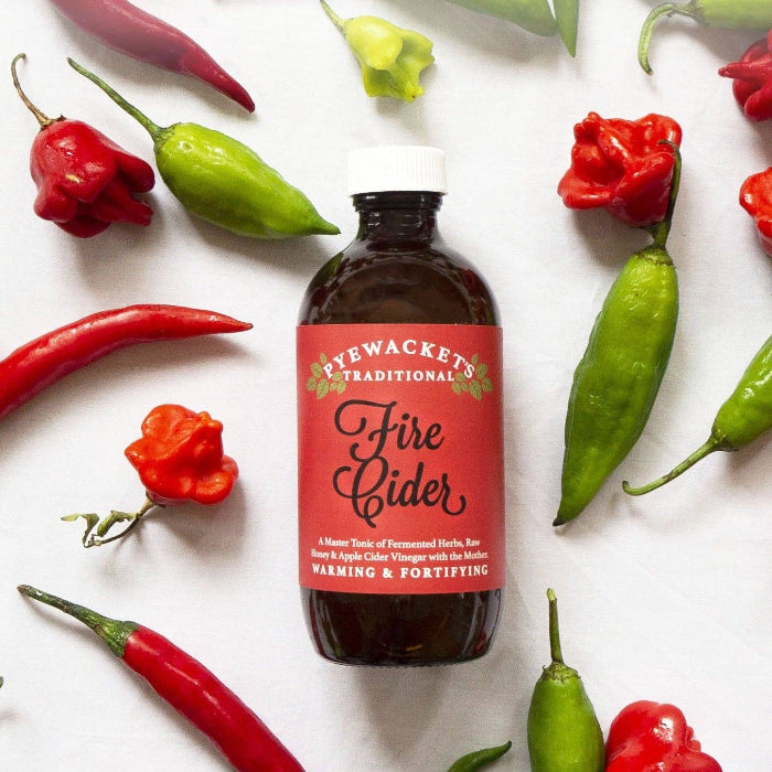 Bottle of Pyewacket's Traditional Fire Cider surrounded by various peppers on a white background