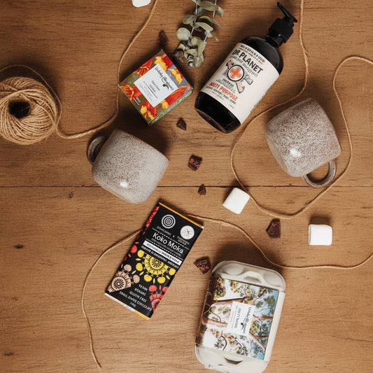 A gift box containing various household items including a bottle of hand wash, living koko chocolate, two ceramic cups, box toilet bombs and premium soap.